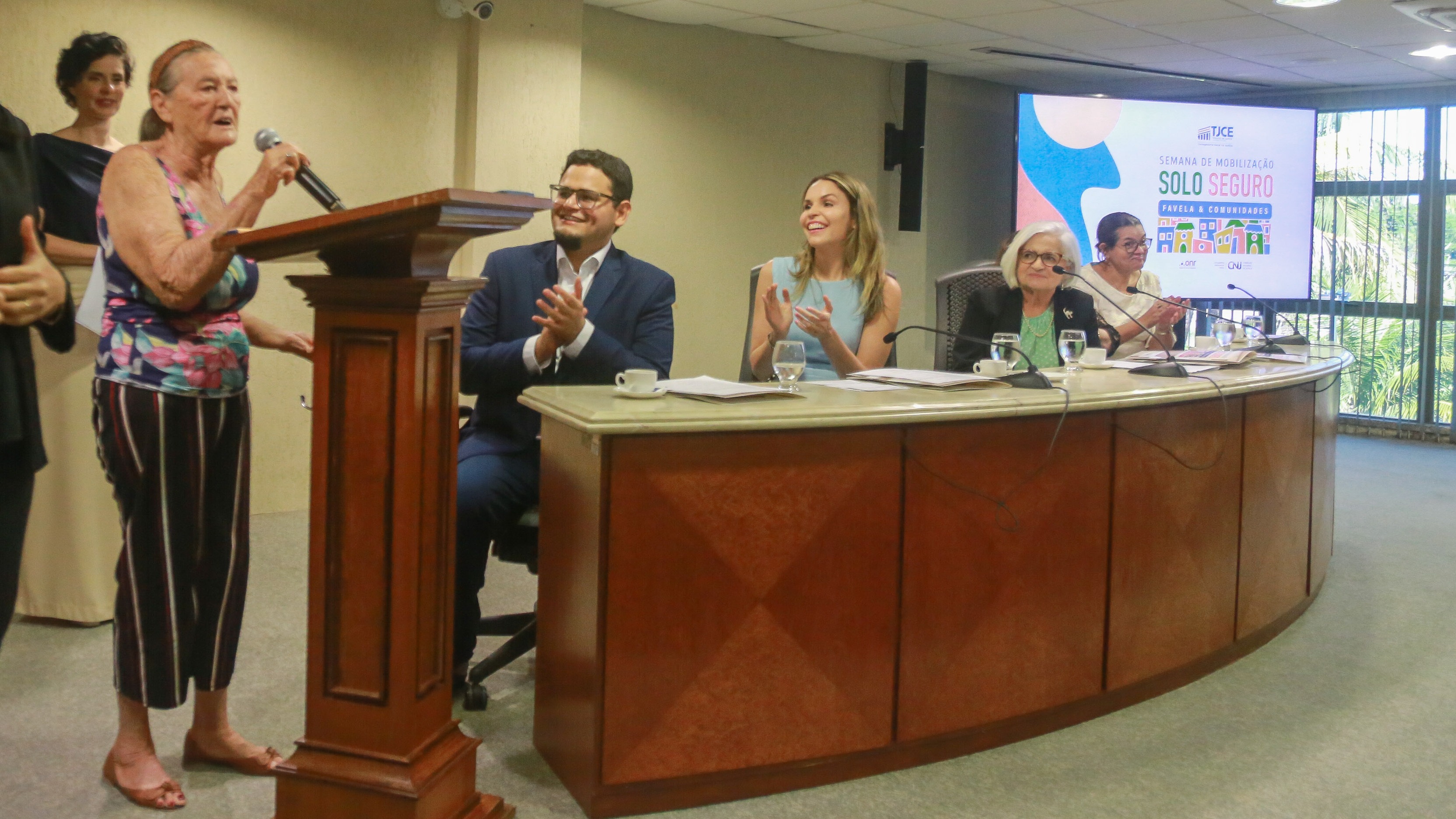 Maria Arcângela de Araújo foi uma das contempladas com o papel da casa. Ela fala ao microfone durante o evento. No palco também está a vice-prefeita, o secretário e os representantes da CGM
